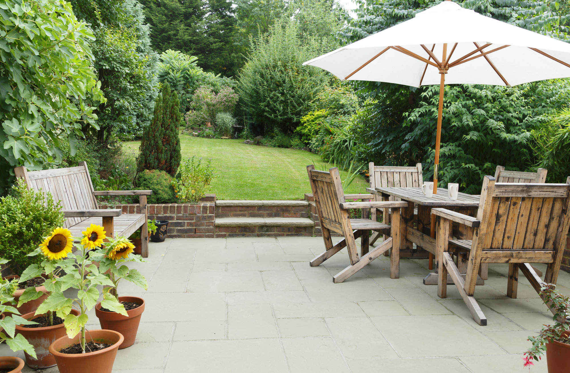 A patio with wooden furniture and a large white umbrella overlooks a lush green garden. Potted sunflowers sit on the stone patio, and dense trees and shrubs surround the grassy lawn in the background.