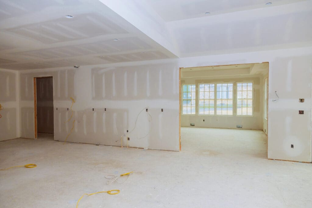 A room under construction with unfinished drywall, exposed electrical wiring, and no flooring. A large open doorway leads to another bright room with multiple windows.
