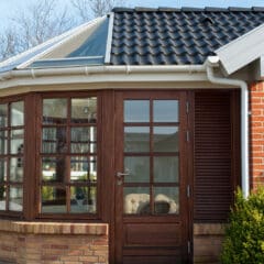 A modern sunroom with large glass windows and a glass door, framed by brown wood and brick walls, attached to a house with a dark tiled roof and white trim; bushes grow in front.