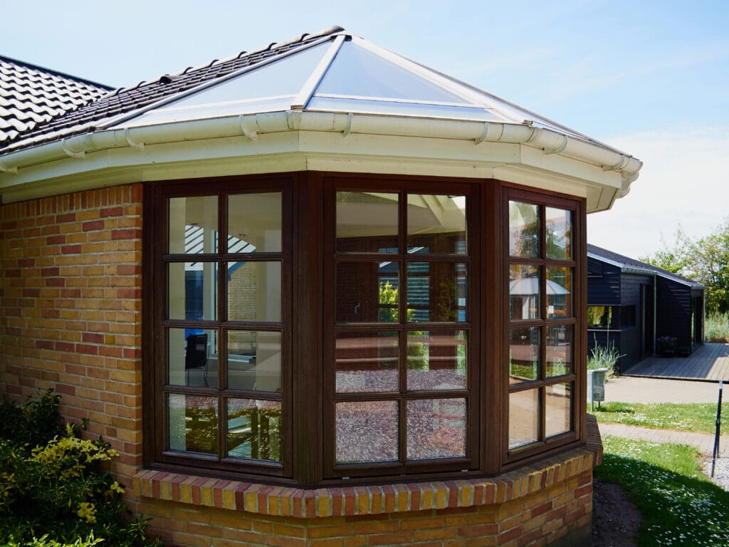 A modern brick house with a large bay window featuring brown frames and multiple glass panes, reflecting the sky and nearby buildings, under a white gutter and a tiled roof.