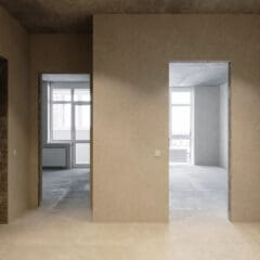 Unfurnished, empty apartment with bare concrete floors and walls, two open doorways, and large windows letting in natural light from outside. Ceiling lights are on.