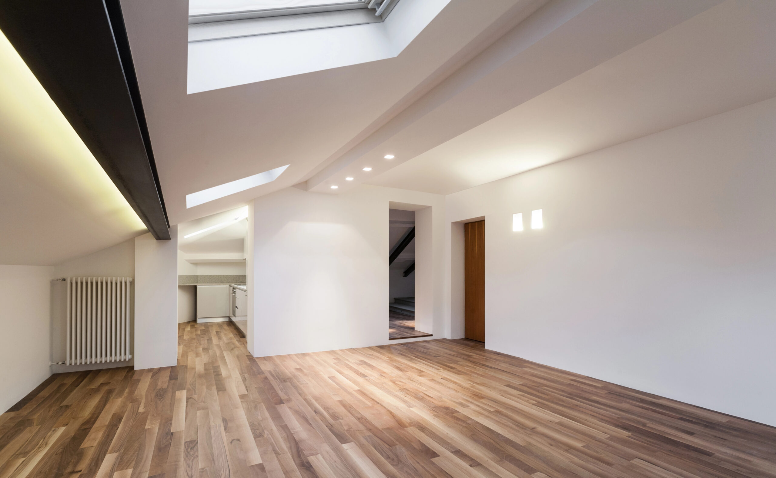 A modern, empty attic room with white walls, wooden flooring, skylights, recessed lighting, and a partial view of a kitchen in the background.