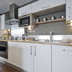 Modern kitchen with white cabinets, stainless steel appliances, a built-in oven, microwave, sink, and stove. Utensils, jars, and a scale are on the counters, with metallic canisters on an open shelf above.