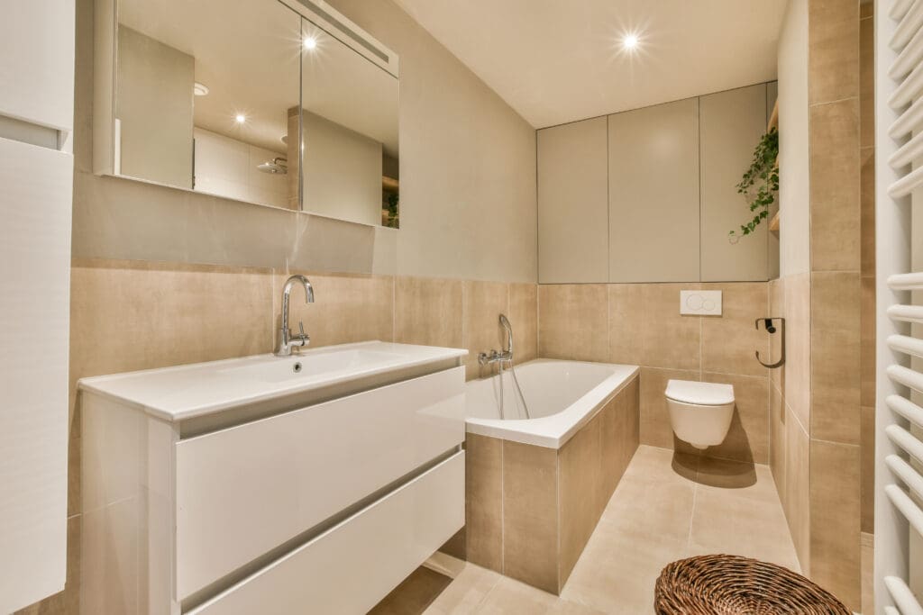 Modern bathroom with beige and white tiles, a wall-mounted sink with drawers, a bathtub, a floating toilet, a large mirror, and a small green plant on a high shelf.