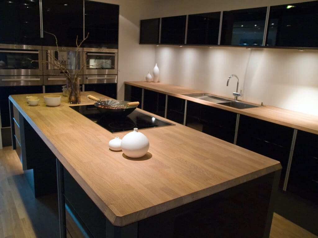 A modern kitchen with black cabinets, wooden countertops, a built-in stovetop on a central island, a sink under wall-mounted cabinets, and minimalist decor including vases and bowls.