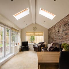 A bright sunroom with skylights, a brick accent wall, leather armchairs, a small TV, a clock above a window, a wooden table with a chair, and large patio doors leading to a fenced garden.
