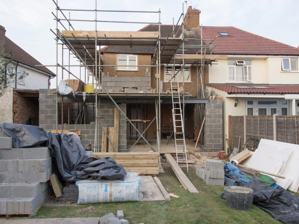 A house under construction with scaffolding and a ladder at the back. Building materials, wooden planks, and bricks are scattered in the garden. The extension's walls are partially built.